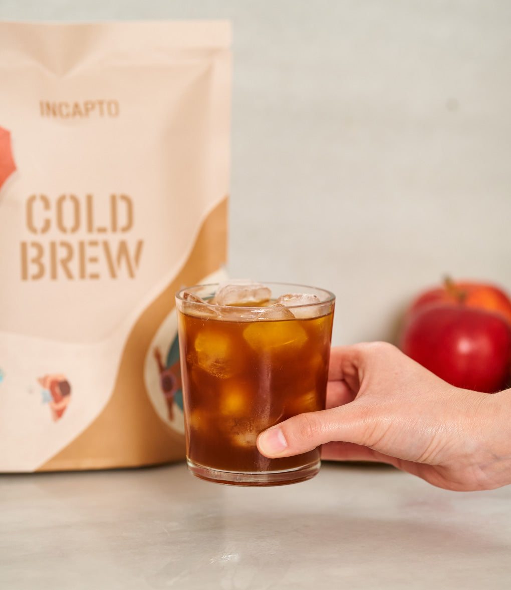 Mano con vaso con cold brew y pack café molido de fondo