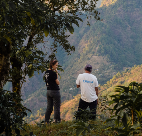 Dos personas de pie en una ladera verde, una con camiseta negra y la otra con camiseta blanca, conversando y mirando hacia un paisaje montañoso en el fondo.