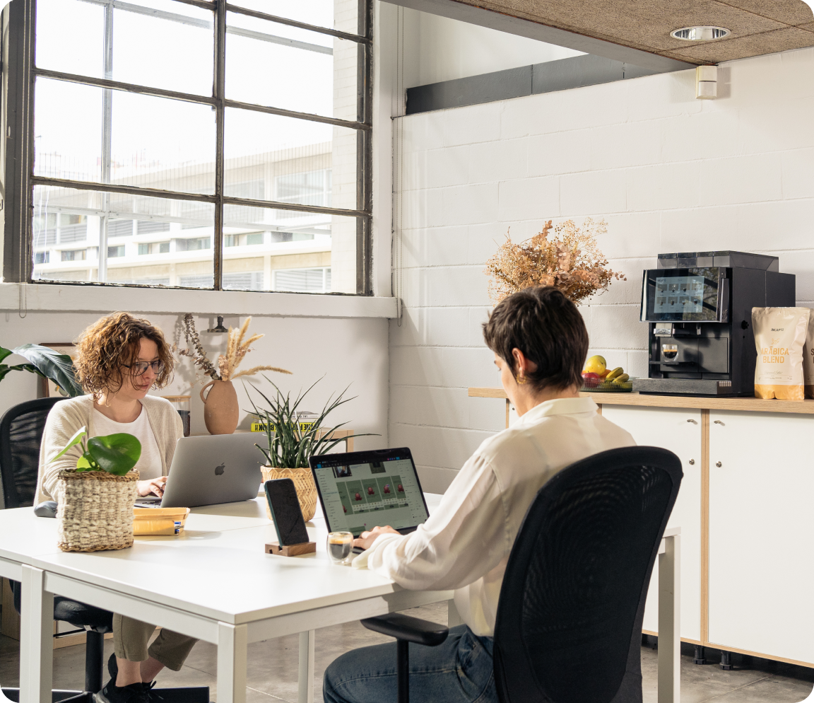 Dos personas trabajando en una oficina INCAPTO luminosa con ventanas grandes, cada una en su computadora portátil. En el fondo, hay una máquina de café junto a una bolsa de café y una canasta de frutas. Plantas y elementos decorativos están distribuidos en el espacio.